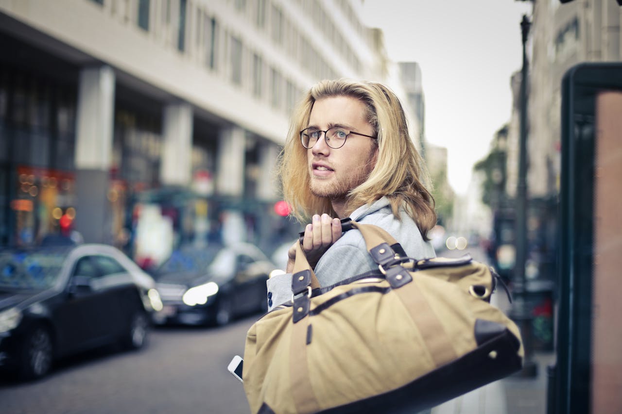 Young man with blonde hair and eyeglasses holding a duffel bag in an urban setting, looking thoughtful.